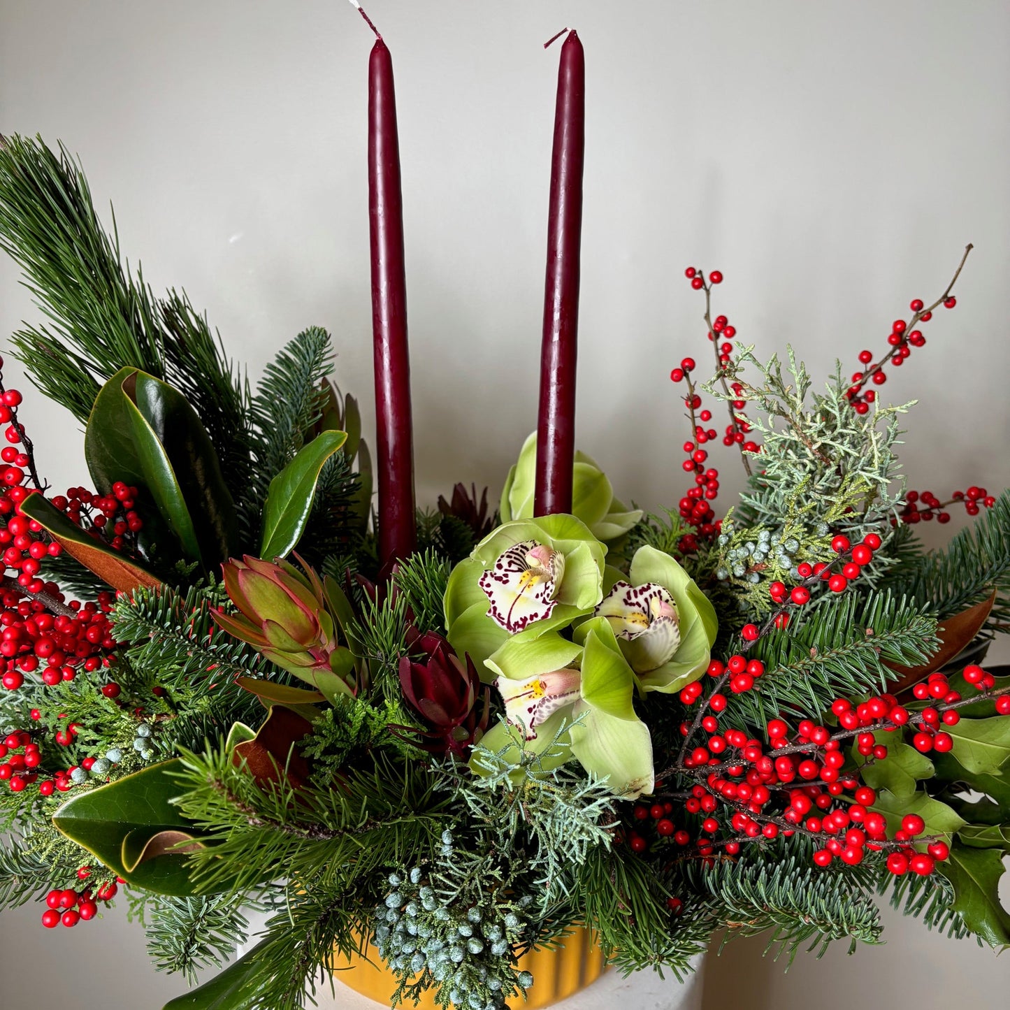 Decorative floral arrangement with green flowers, red berries, and candles on a white surface.