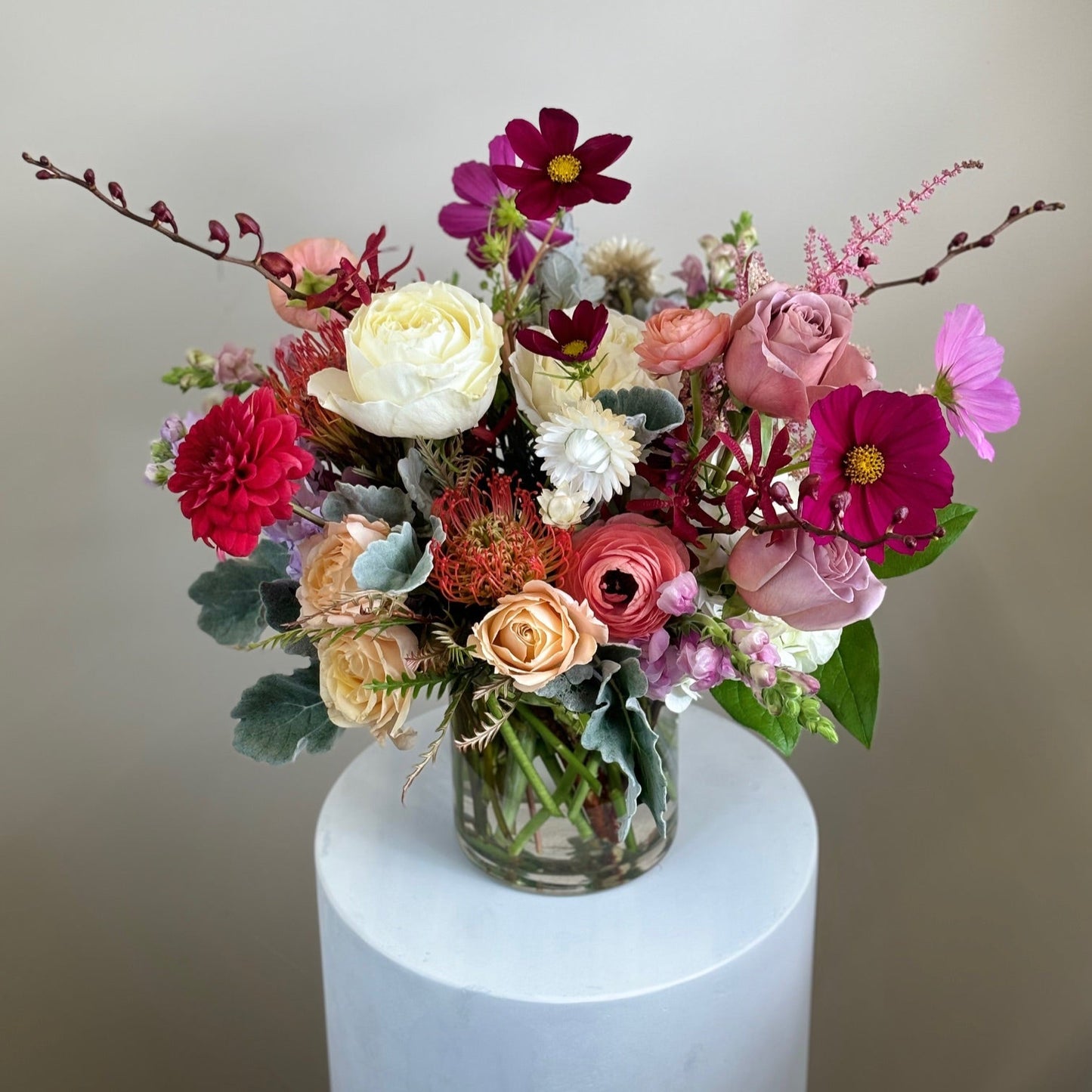 A colorful flower arrangement featuring roses, peonies, and calla lilies in a clear vase, displayed on a white pedestal.