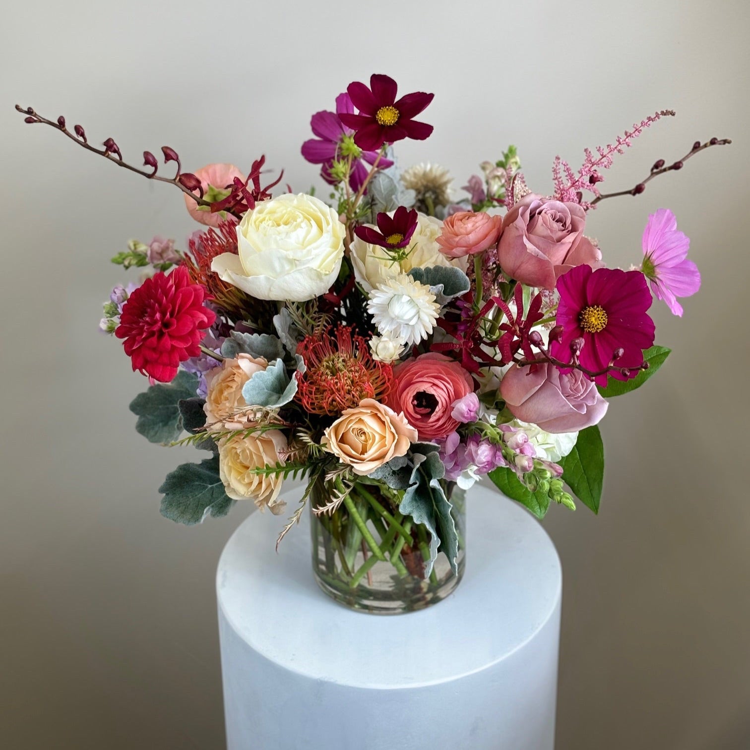 A colorful flower arrangement featuring roses, peonies, and calla lilies in a clear vase, displayed on a white pedestal.