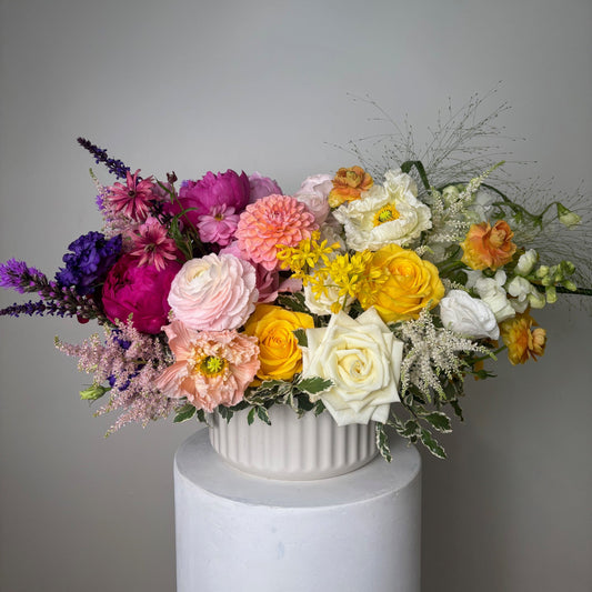 Colorful flower arrangement spilling out of a white cylindrical container on a gray background