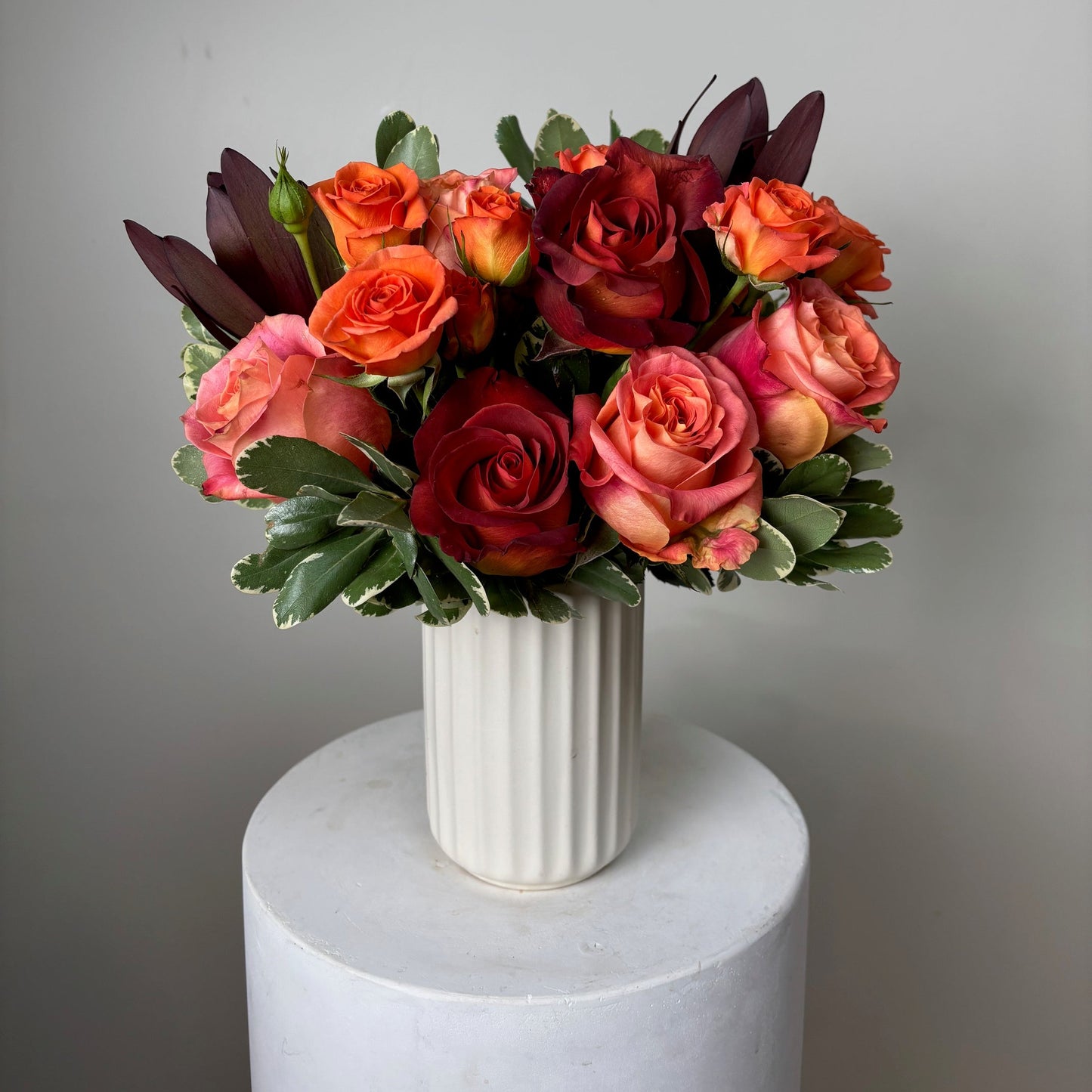 Bouquet of red, orange, and pink roses in a white vase on a light gray background