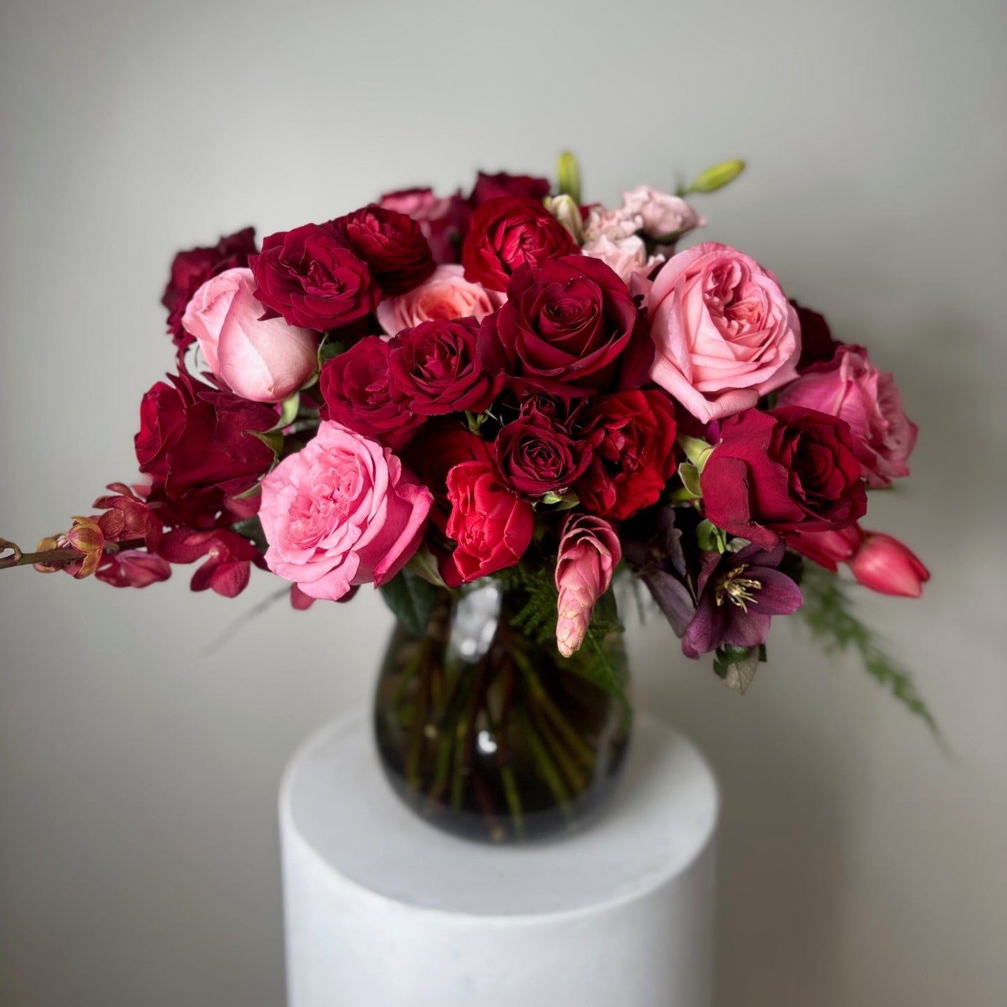 Bouquet of red and pink roses in a clear vase on a white pedestal against a gray background